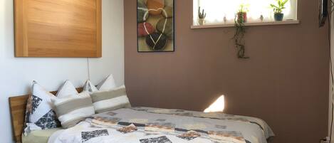 Ein Schlafzimmer mit einem Bett, einem Holz-Kopfstück, einem Fenster mit Pflanzen und einem an der Wand befestigten Kunstwerk.