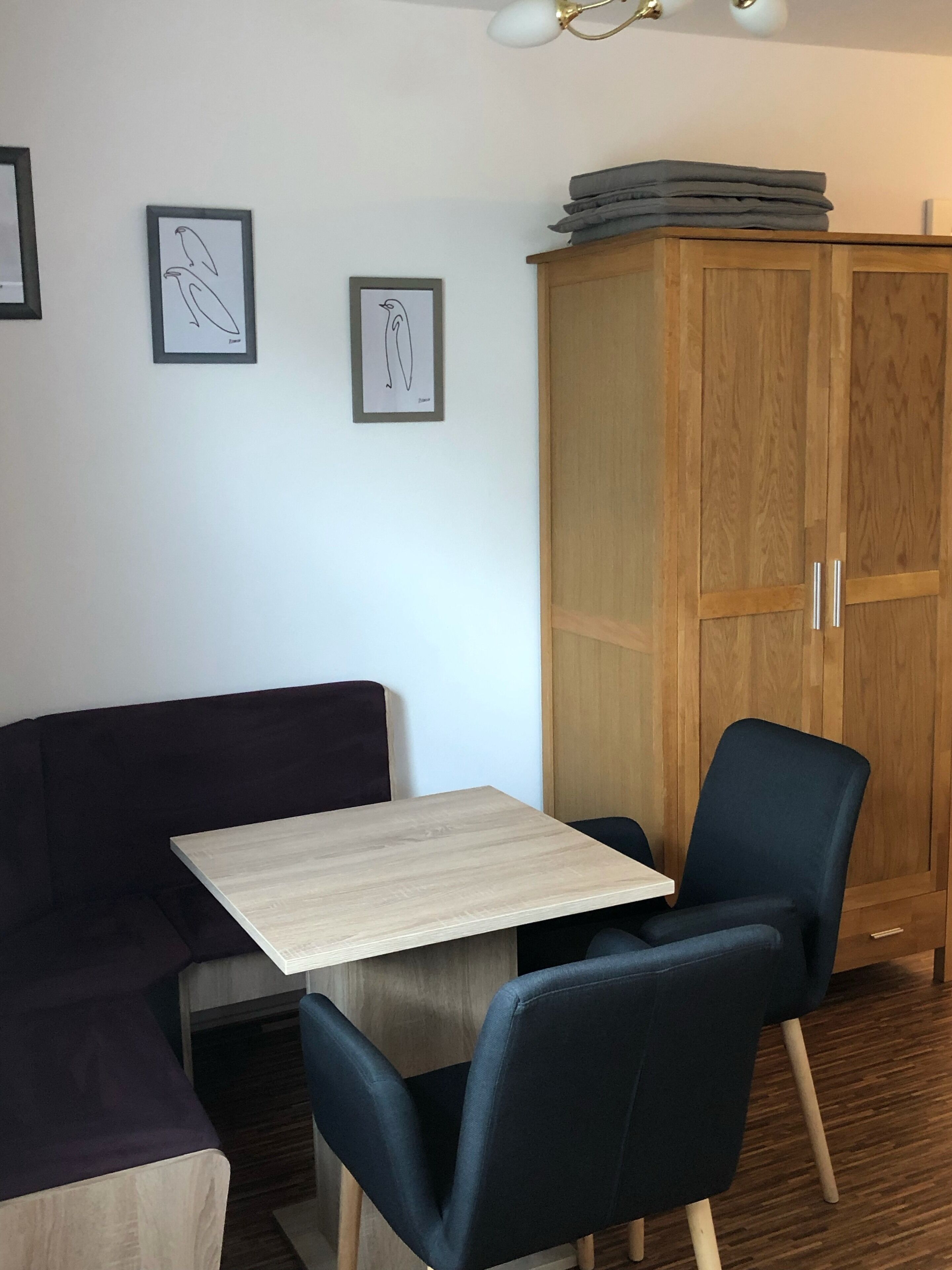Une pièce comprenant un meuble en bois, une chaise bleu foncé, une table de couleur claire et deux œuvres d’art encadrées accrochées au mur.