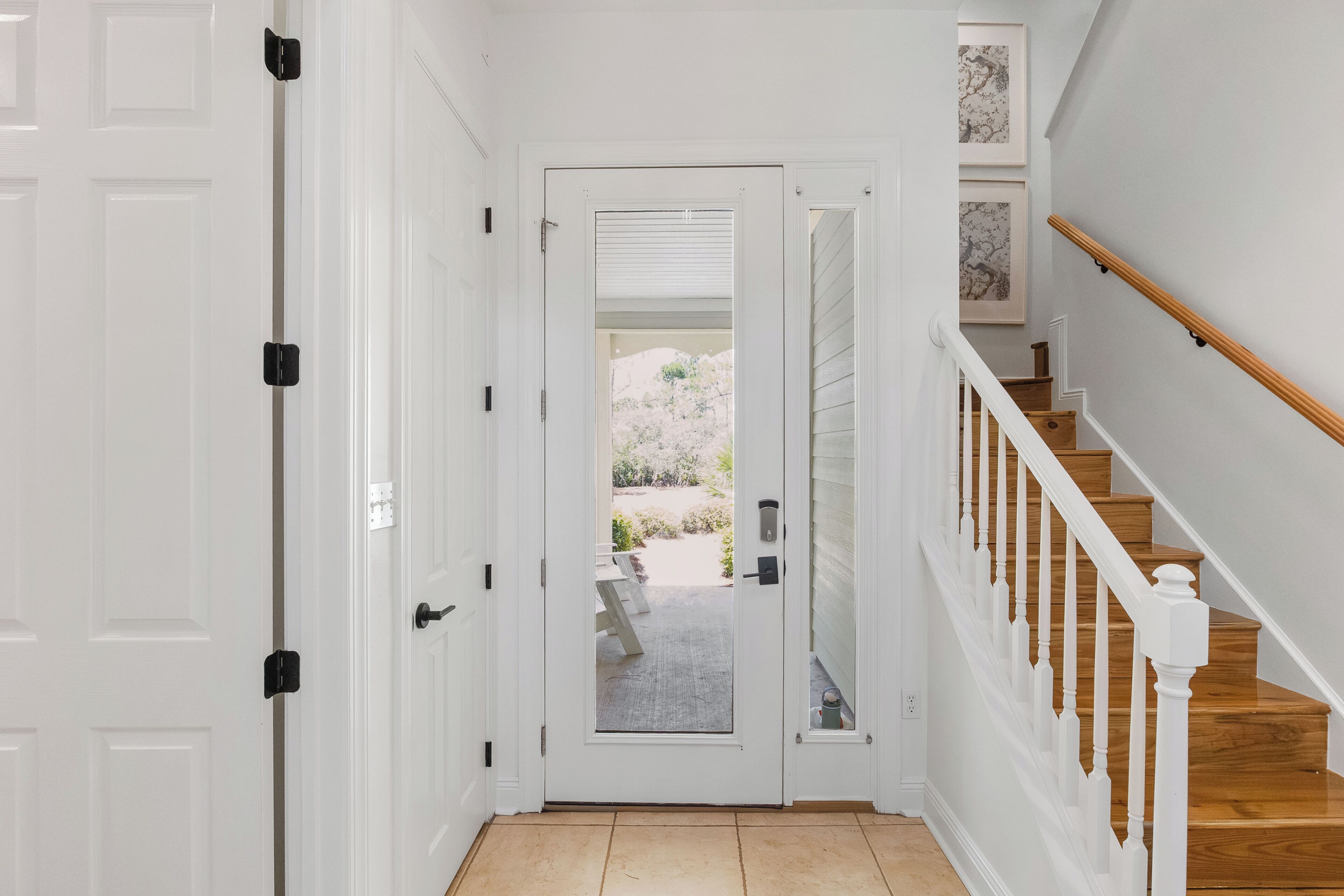 Un pasillo con una puerta blanca, una escalera con escalones de madera y barandillas blancas, y vistas a un área exterior con plantas.