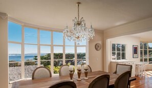Un espace repas comprenant une grande table, des chaises et un lustre. De grandes fenêtres offrent une vue sur l’océan et les arbres.