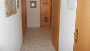 Un couloir avec un sol carrelé, une porte en bois et une petite poubelle.