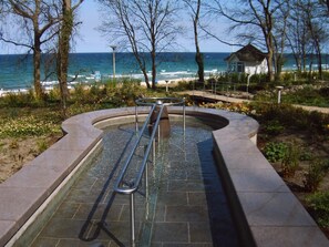 A swimming pool with a metal ladder, surrounded by a stone deck, with a view of the ocean and beach in the background.