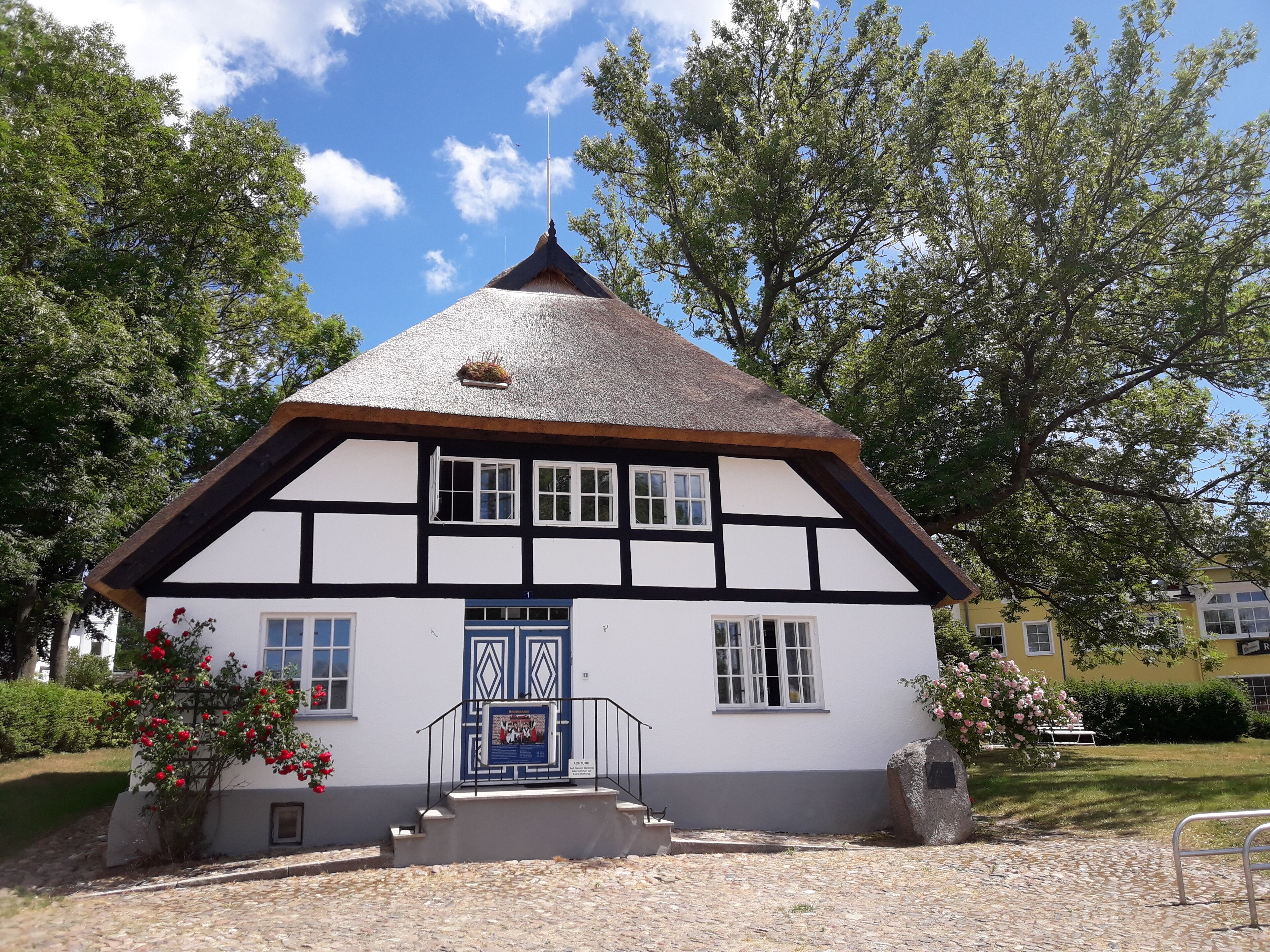 Ein traditionelles Fachwerkhäuschen mit einem steilen Walmdach, einem zentralen Eingang mit einer blauen Tür und einer kleinen Veranda mit Stufen.
