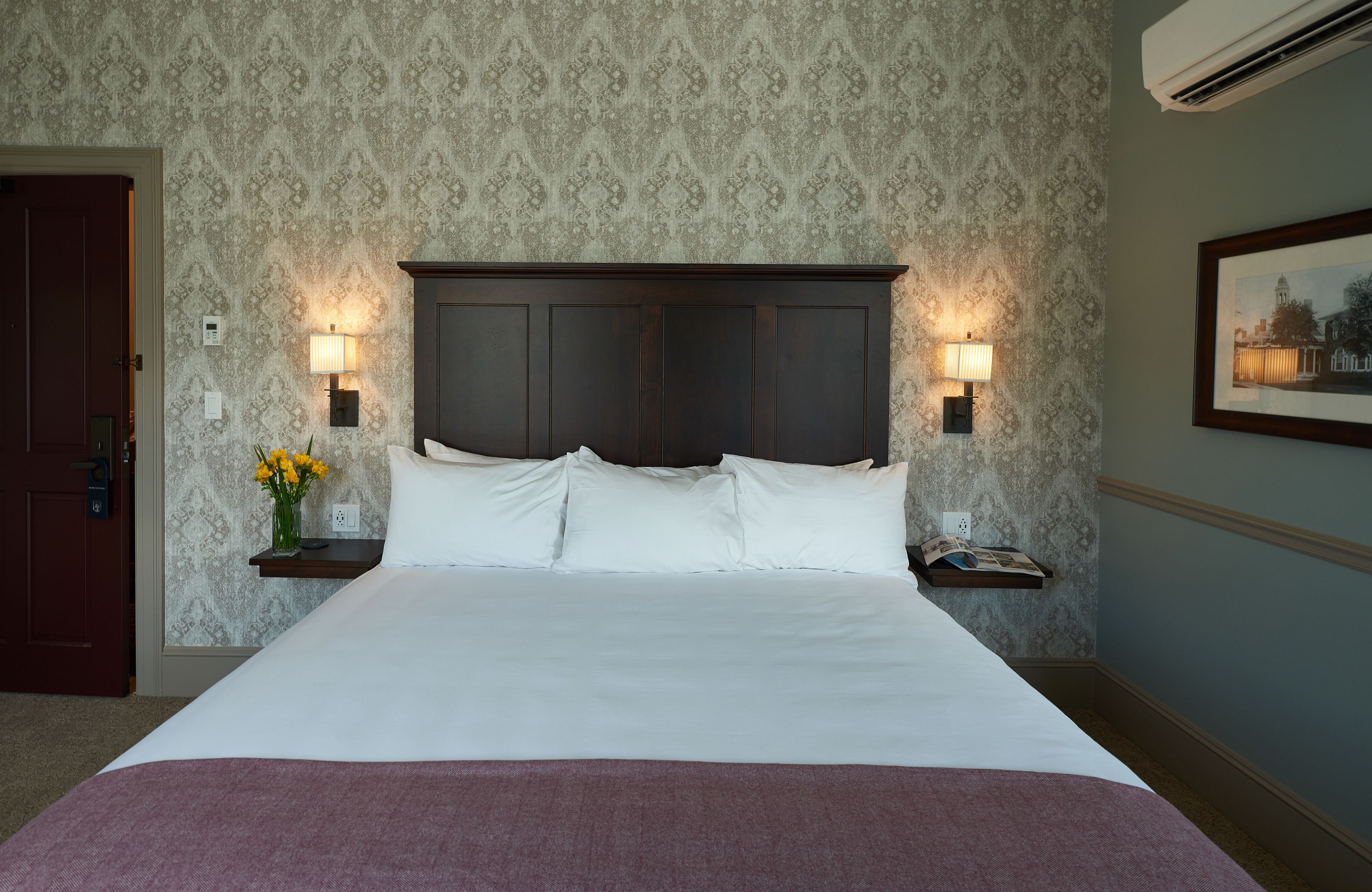 A neatly made bed with white linens and a dark headboard, flanked by two bedside lamps and a small table with a vase of flowers.