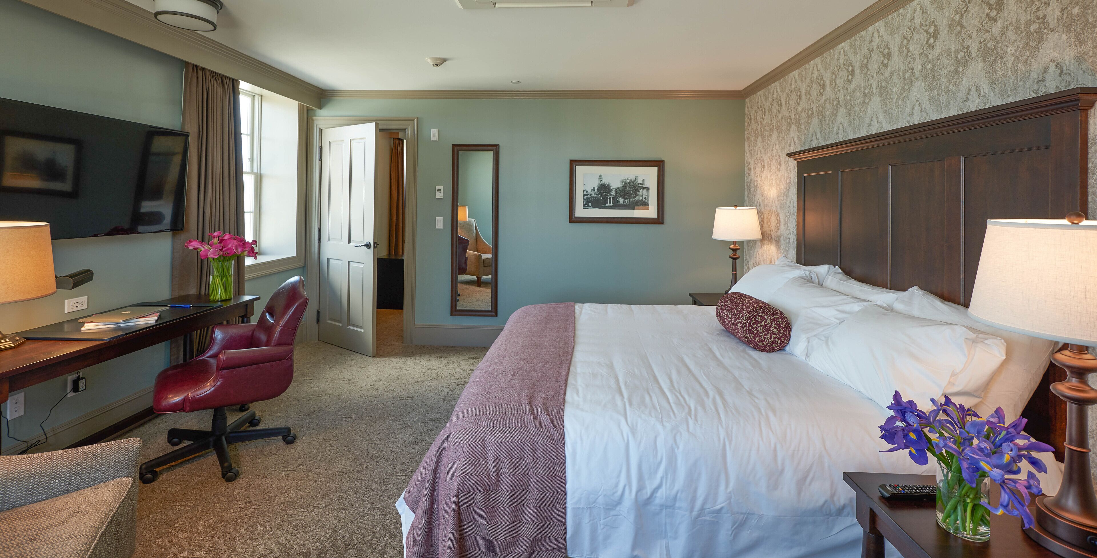 Una habitación de hotel con cama, escritorio, silla, televisor, lámparas y un florero con flores.