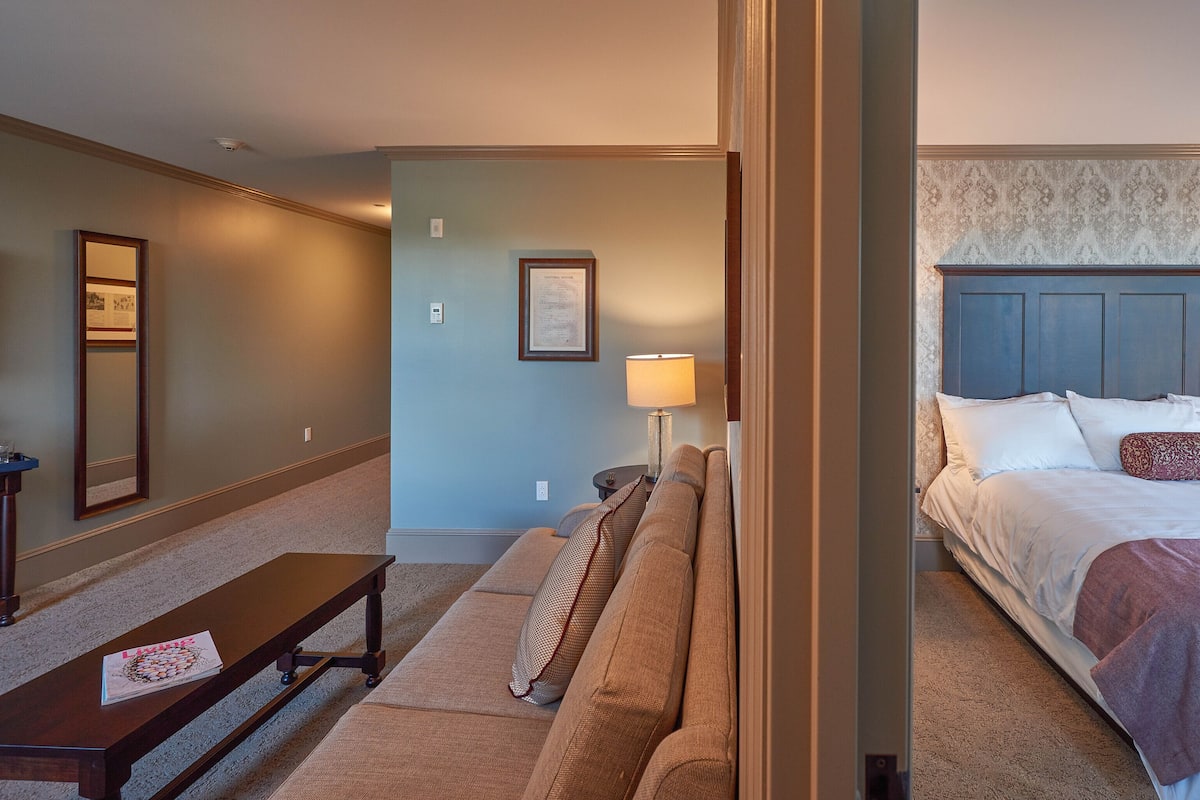 A hotel room with a bed, a sofa, a bench, a lamp, and a framed certificate on the wall.