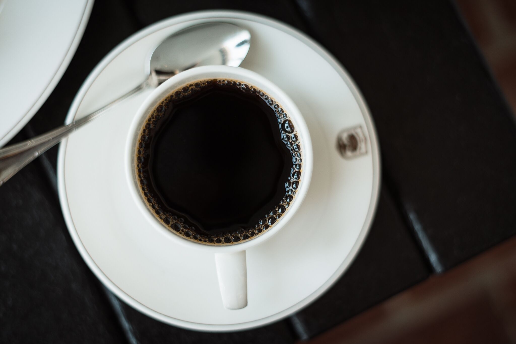 A white cup of black coffee on a white saucer with a spoon beside it, placed on a dark surface.
