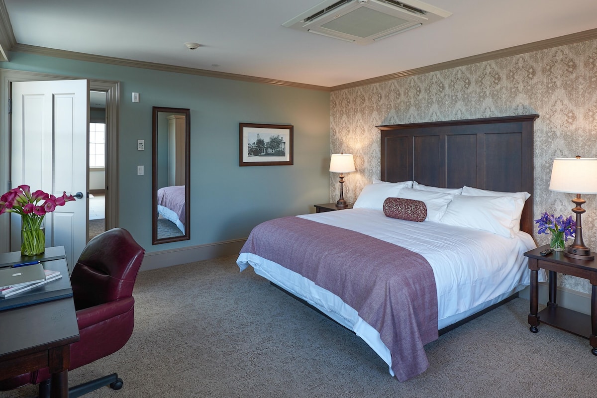 A hotel room with a bed, bedside tables, a desk, a chair, a mirror, and a picture on the wall.