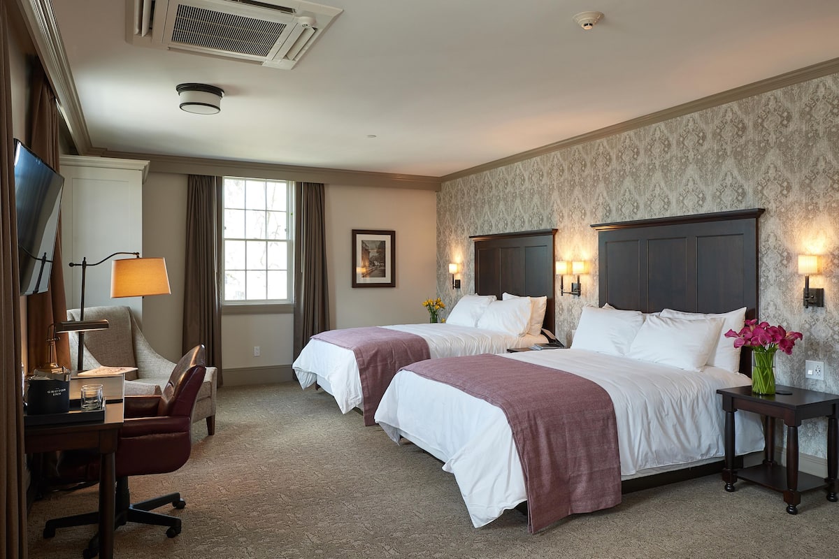 A hotel room with two beds, a desk, a chair, a TV, and a window with curtains.