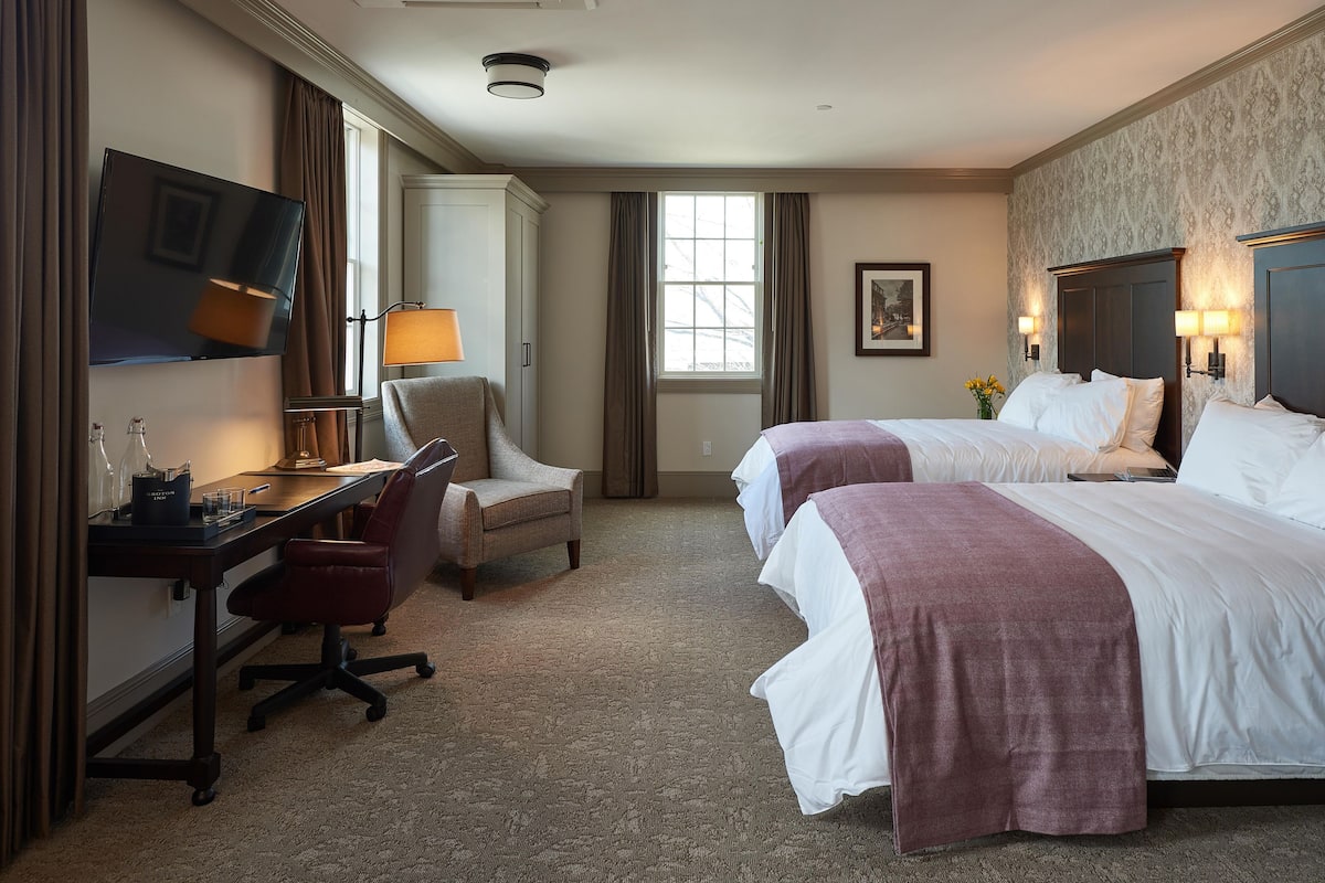 A hotel room with two beds, a desk, a chair, a TV, and a window.