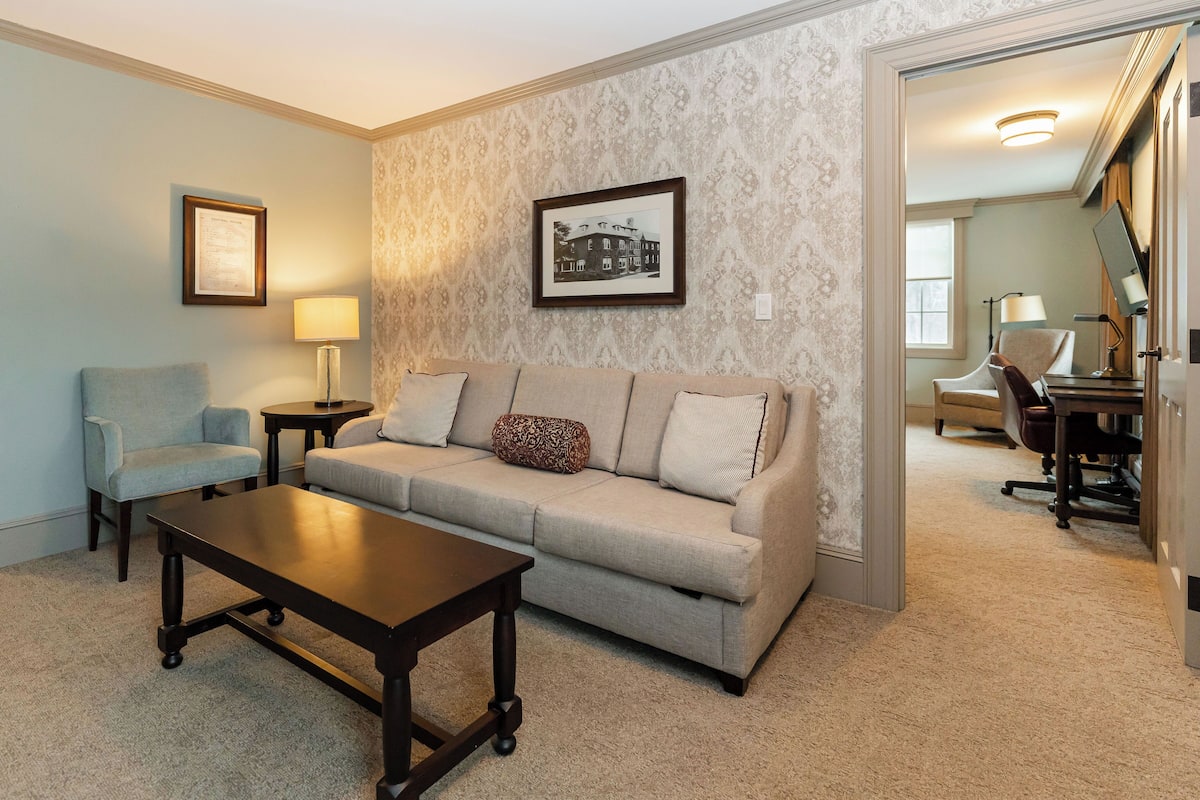 A living room with a sofa, a small table, a chair, and a lamp. There is a framed picture on the wall and a staircase leading to another room.