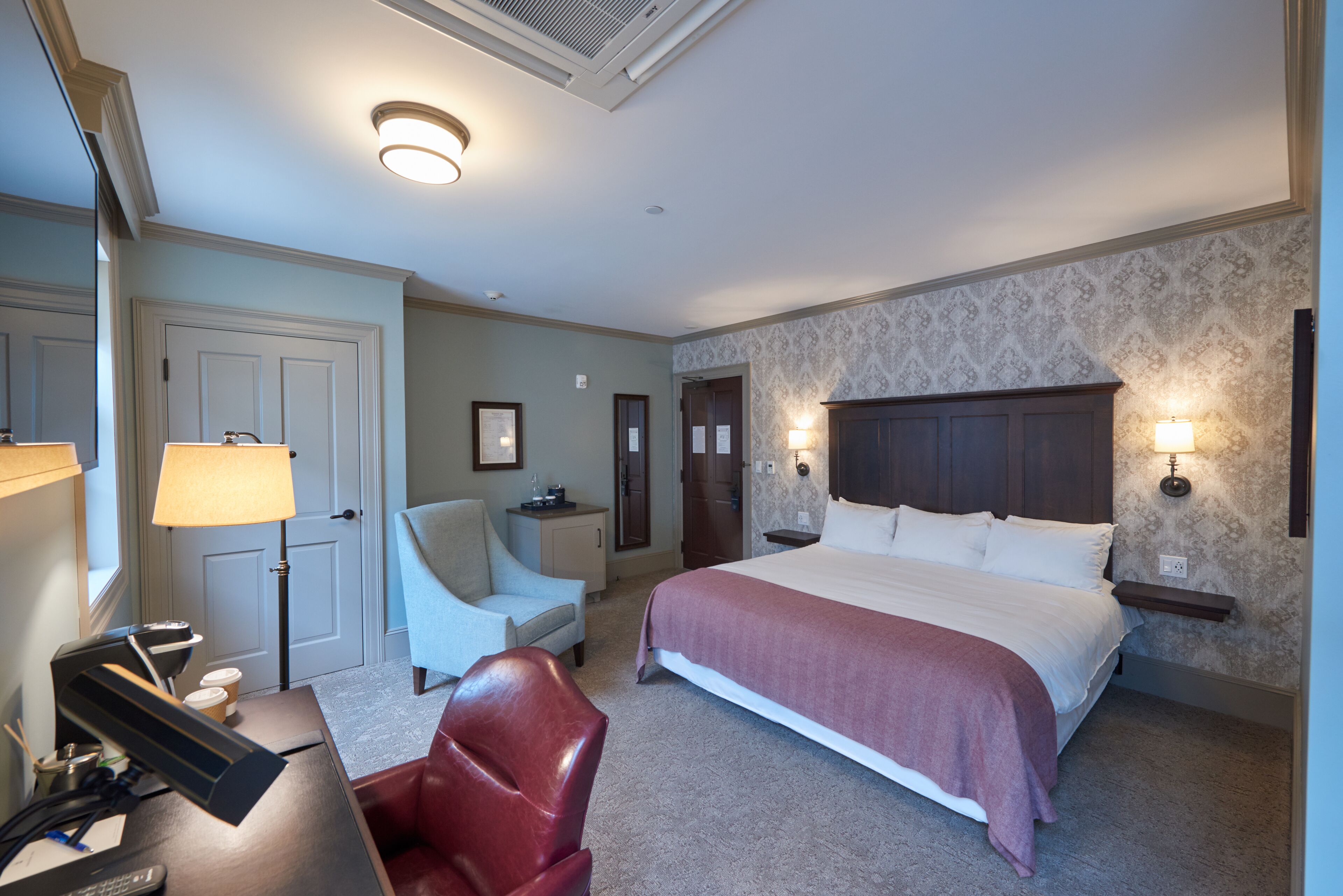 A hotel room with a large bed, a desk with a lamp, a chair, and a desk lamp.