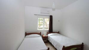 Een kleine, schone kamer met twee eenpersoonsbedden, een nachtkastje, een raam met gordijnen en een airconditioning.