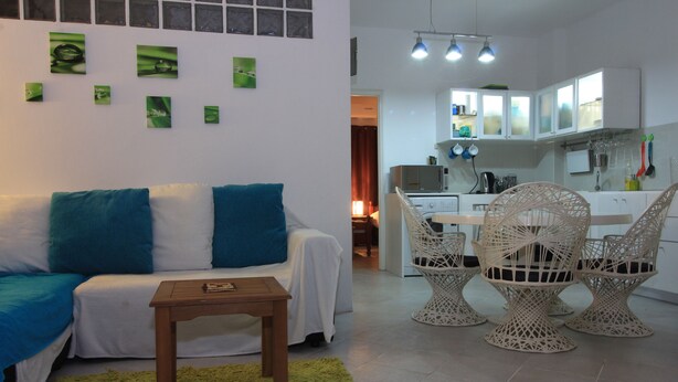 Een woonkamer met een bank, een salontafel en rieten stoelen. Achtergrond toont een keuken.