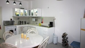 Een kleine keuken met witte kastjes, een magnetron, koffiezetapparaat en een spoelbak. Boven het aanrecht hangen drie hanglampen en er staat een witte eettafel met stoelen.