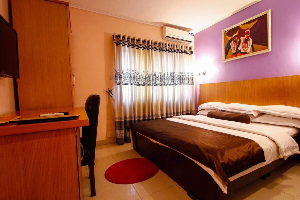 Quarto de hotel com cama, escrivaninha, cadeira, tapete vermelho e um quadro na parede.