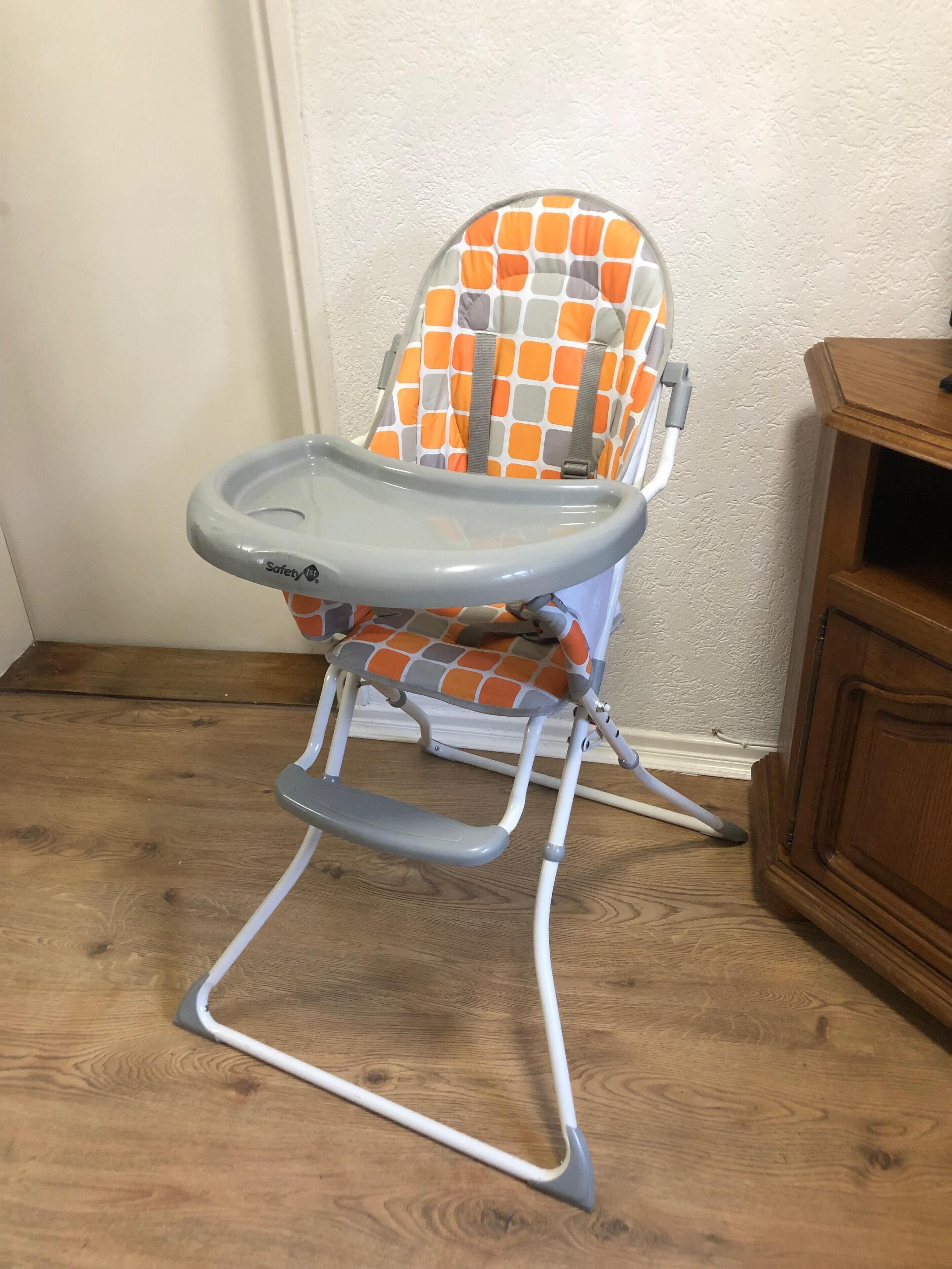 A high chair with a safety strap and a tray, placed on a wooden floor against a white wall.