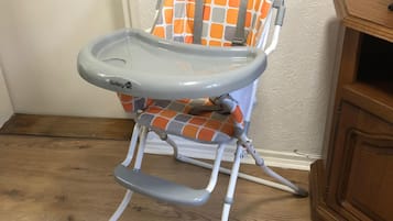 A high chair with a safety strap and a tray, placed on a wooden floor against a white wall.
