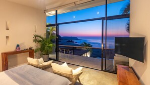 A bedroom with a large window overlooking a beach at sunset, a bed, a TV, and a small seating area.
