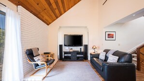 A modern living room with a wooden ceiling, a flat-screen TV, a black leather sofa, a black leather chair, and a small table with a lamp.