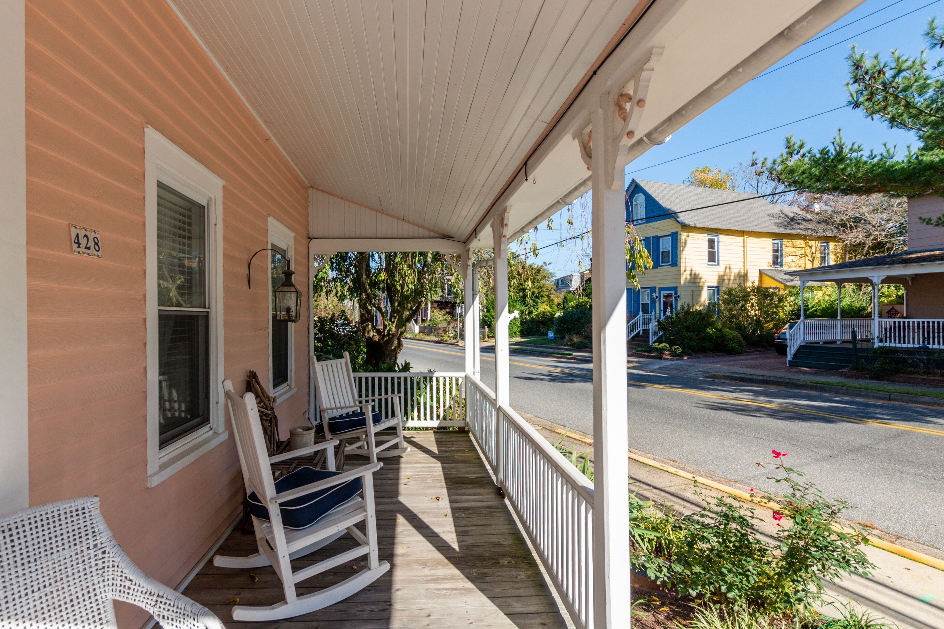 A porch with rocking chairs and a white chair, overlooking a street with houses and a clear sky.