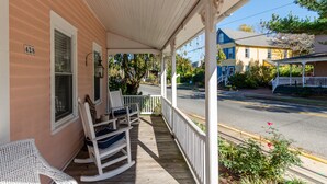 Eine Veranda mit Schaukelstühlen und einem weißen Stuhl, mit Blick auf eine Straße mit Häusern und blauem Himmel.