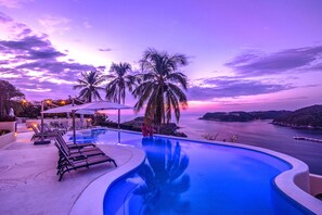 Ein Pool mit Liegestühlen und Sonnenschirmen mit Blick auf ein Gewässer und Palmen bei Sonnenuntergang.