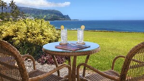 Une table en osier avec deux verres contenant une boisson, un livre, et une vue sur l’océan et les montagnes.