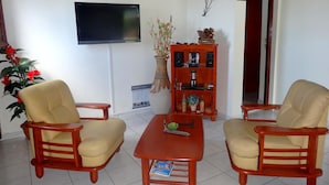 A living room with two beige armchairs, a red coffee table, a TV on the wall, and a wooden cabinet with decorative items.