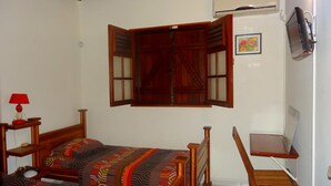 A bedroom with a bed, a desk, and a chair. There is a window with wooden shutters and an air conditioning unit on the wall.