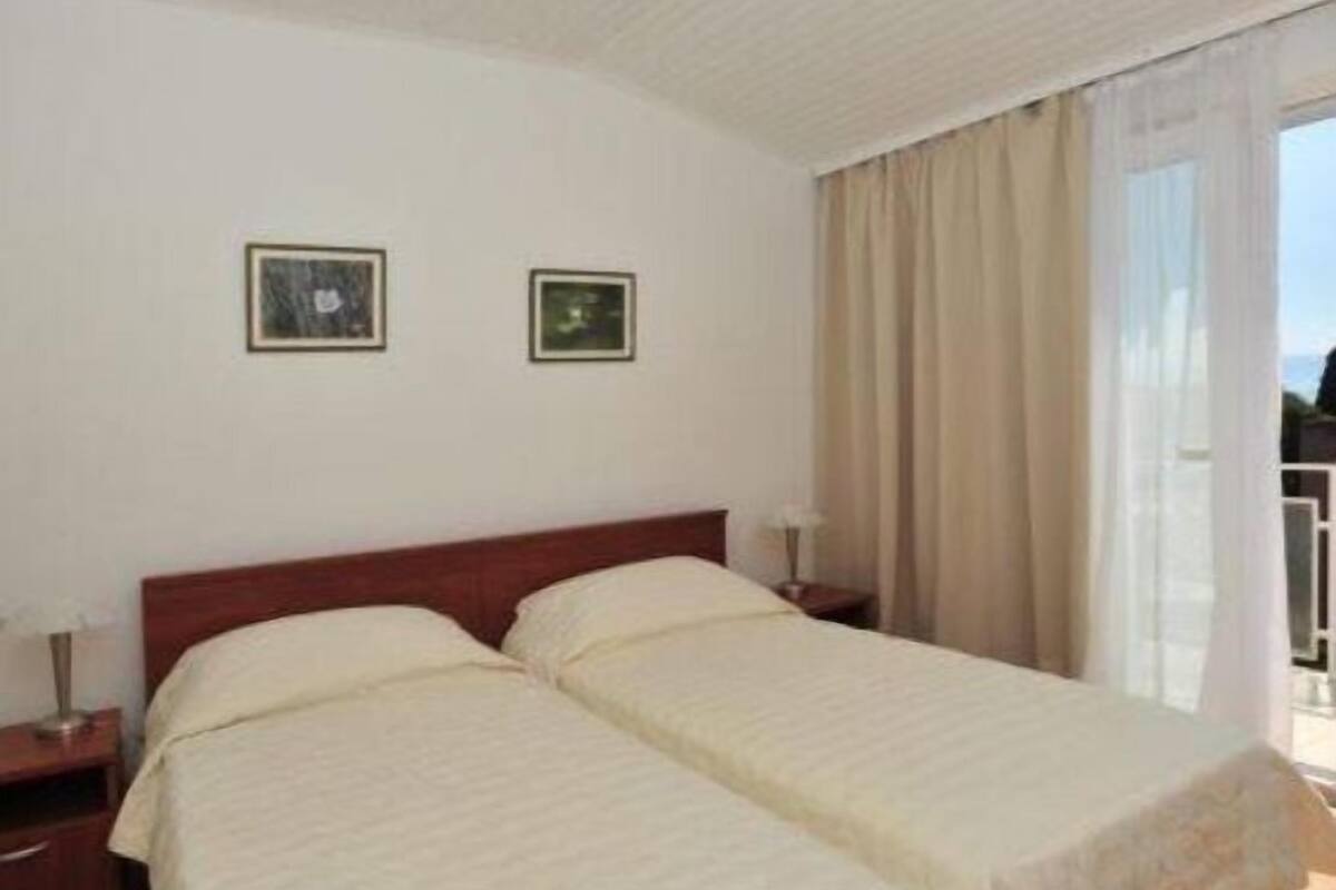 Habitación de hotel con dos camas individuales, cabecero de madera, mesita de noche con lámpara y ventana con cortinas.