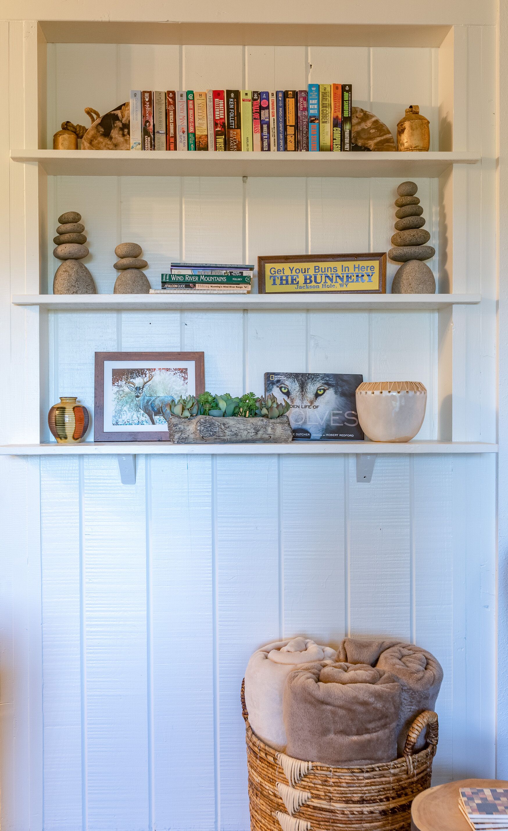 Une bibliothèque avec des livres et des objets décoratifs, un tableau encadré et un petit panier contenant un coussin.