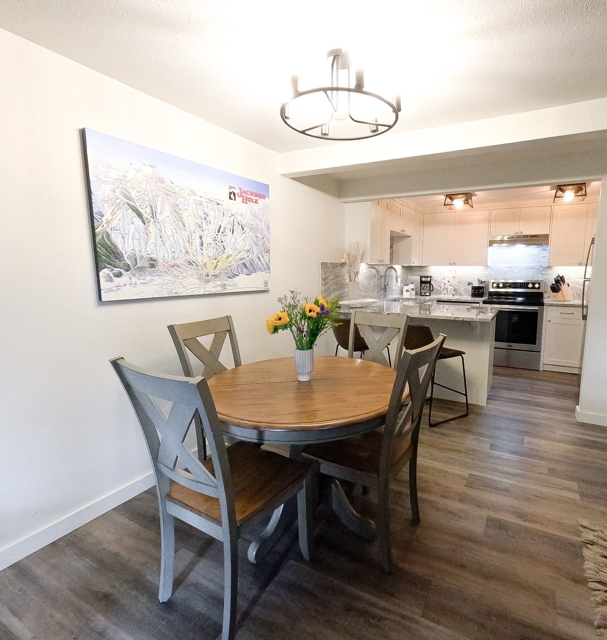 Un comedor con una mesa redonda de madera y sillas, un jarrón con flores y una pintura de Jackson Hole en la pared.