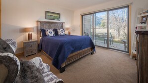 Un dormitorio con una cama grande, una mesita de noche, una lámpara, una estantería y un ventanal corredero que da a un patio con mobiliario exterior.