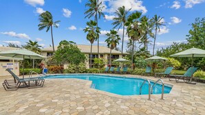 A pool area with lounge chairs and umbrellas, surrounded by palm trees and a building with a sign that reads "HOTEL".
