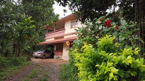Une maison à deux étages avec un balcon, entourée d’une végétation luxuriante et une voiture garée dans l’allée.