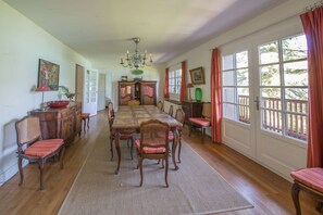 A dining room with a long wooden table, red cushioned chairs, a chandelier, and large windows with curtains.