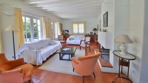 Ein geräumiges Wohnzimmer mit einer Sofagarnitur, einem Couchtisch und einem Sessel. Es gibt Fenster mit Vorhängen, eine Lampe und ein Gemälde an der Wand.