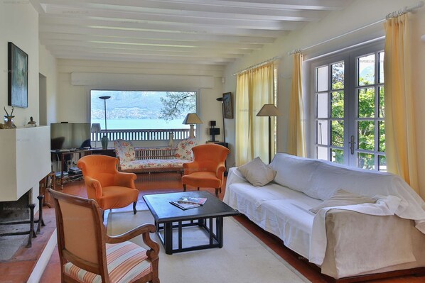 A living room with a sofa, two armchairs, a coffee table, and a television. There is a balcony with a railing and a view of the outside.