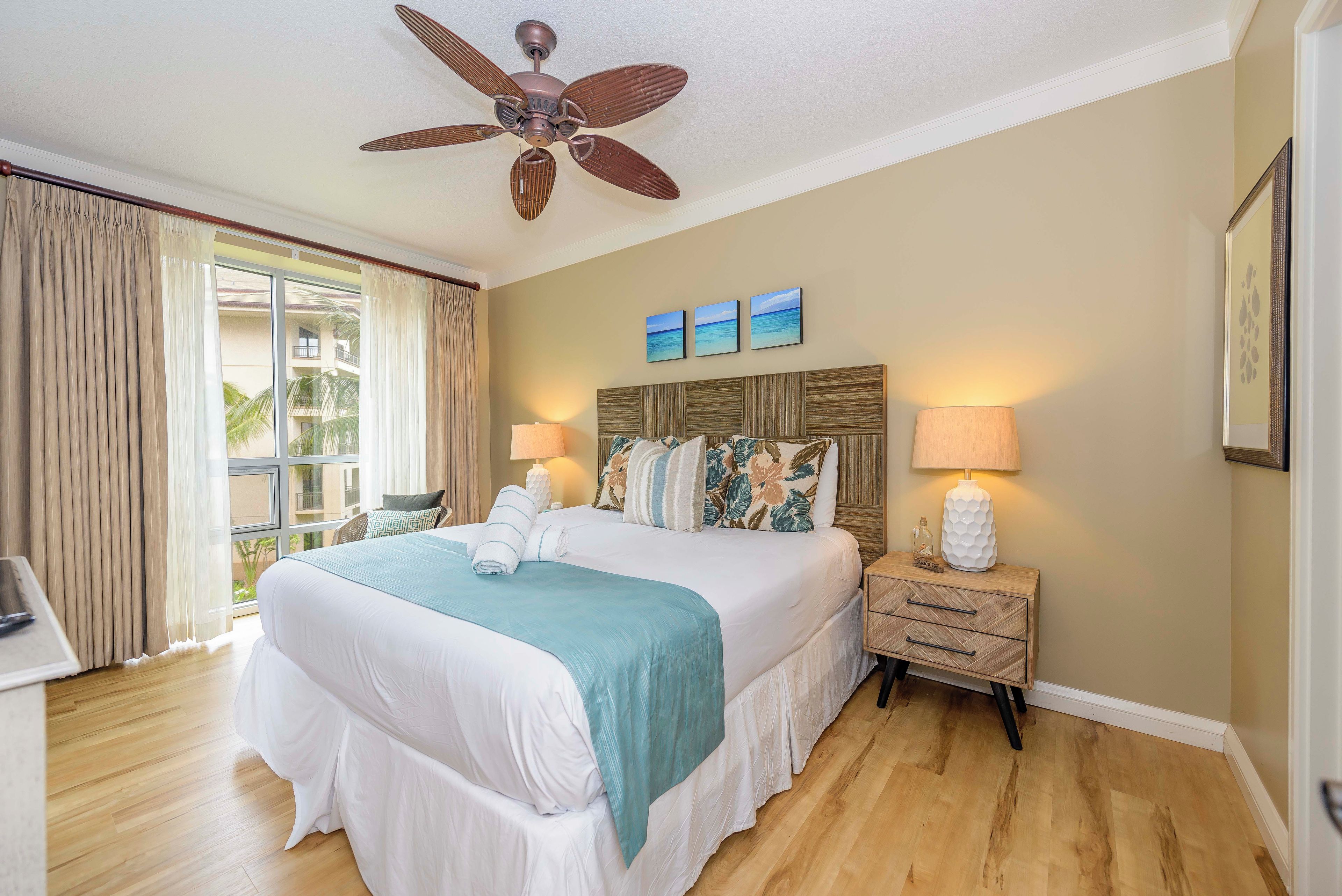 A hotel room with a large bed, a ceiling fan, a nightstand with a lamp, and a view of palm trees through the window.
