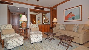 A living room with a sofa, two armchairs, a coffee table, and a side table with lamps. There is a framed picture on the wall and a wooden door in the background.