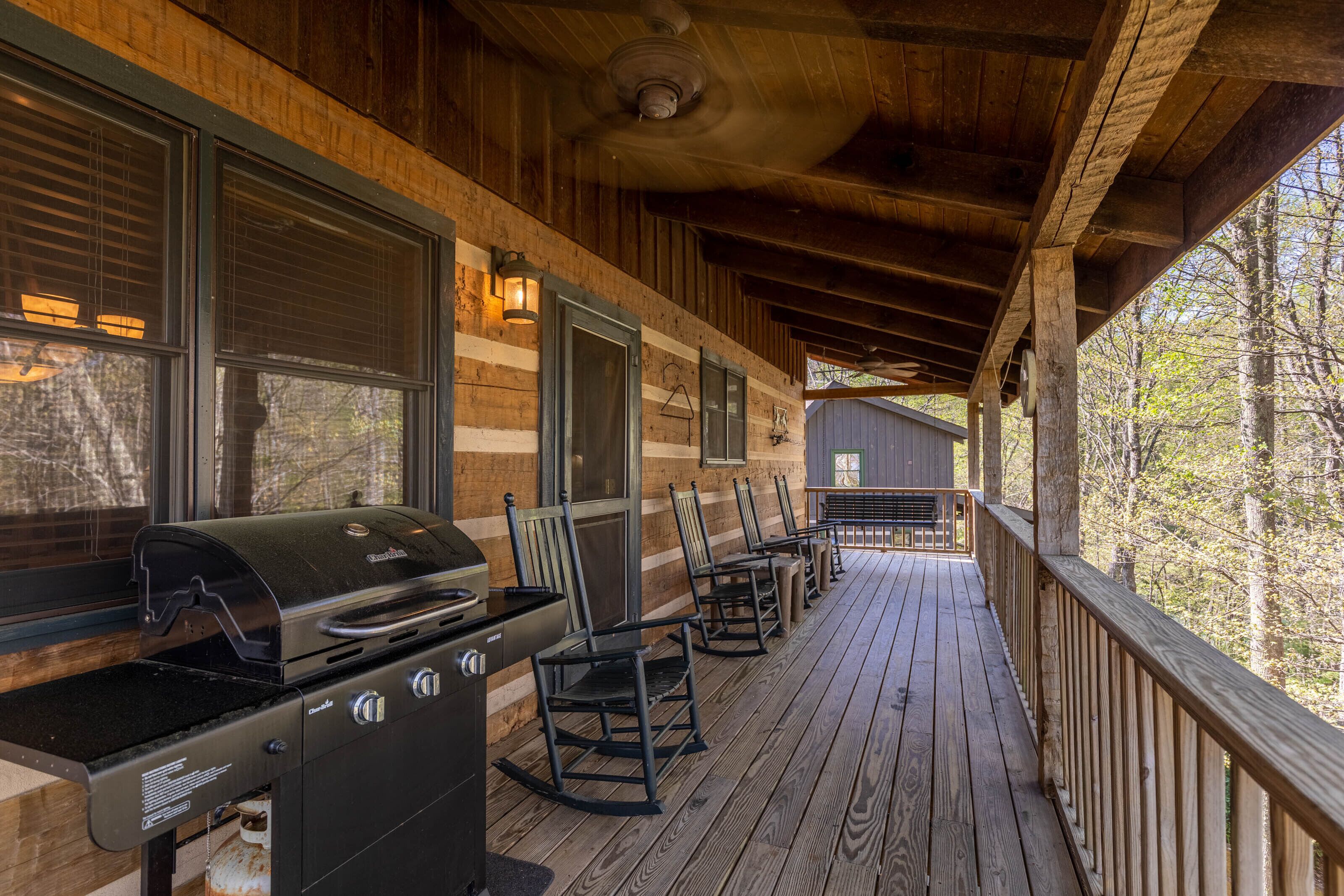A covered outdoor deck with a barbecue grill, wooden chairs, and a view of trees.