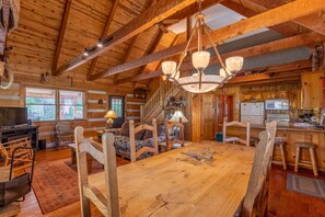 A rustic cabin interior with a wooden dining table, chairs, a staircase, and a kitchen area.