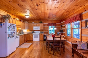 A cabin interior with wooden walls, a kitchen area, a dining table with chairs, and a window with a view of trees.