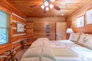 A wooden cabin interior with a bed, a chair, a ceiling fan, and framed botanical prints on the walls.