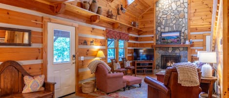A cozy log cabin interior with a stone fireplace, wooden furniture, and a television.
