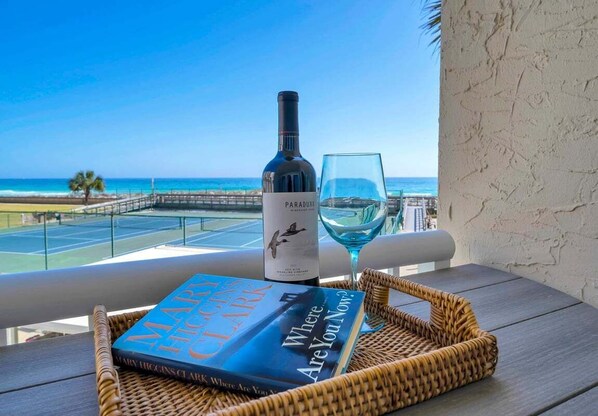A balcony with a view of the beach, a bottle of wine, a glass, and a book.