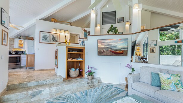 A spacious living area with a staircase leading to an upper level, a kitchen in the background, and a large window offering a view of the outdoors. - Pali Uli 8, The Gem, honeymoon or romantic getaway (Princeville)