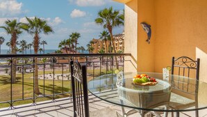 Un balcón con una mesa y silla de vidrio, con vistas a una zona de piscina con palmeras y edificios.