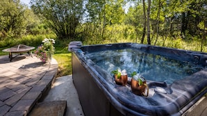 Ein Whirlpool mit zwei Kupferbechern, gefüllt mit Getränken, umgeben von einer Holzdielenveranda und einer Rasenfläche mit Bäumen.